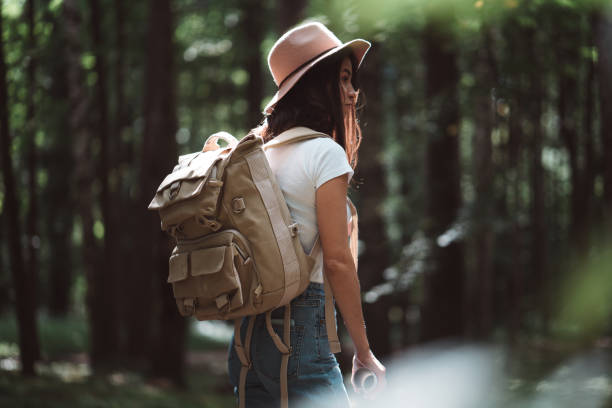 istock Back view on cute young woman with hat, backpack and location map in hand among trees in forest at sunset. 1013816914