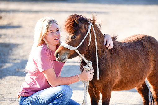 This is a color photograph of a American teenage girl with her miniature pony in Spanish Fork, Utah, a western USA state. She has long blonde hair and hugs the small horse on a spring afternoon.