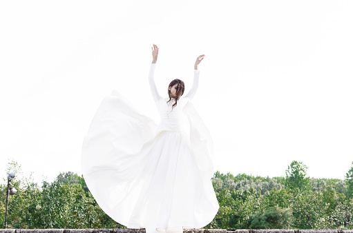 Beautiful young woman is posing on wind in nature in long white dress.