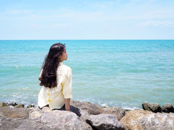 istock young girl sitting on the rocks at beach. 1013964350