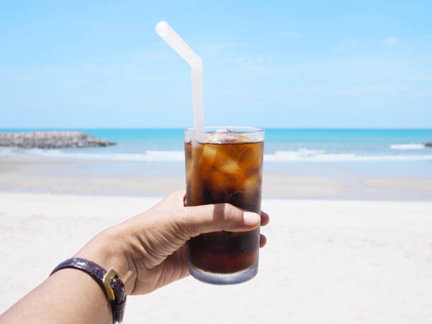 Close up hand holding glass of soft drink with ice over summer beach background.