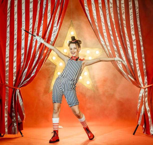 Funny Magician Girl at Circus Show Beautiful young girl dressed in a magician costume shows the circus performance. The preadolescent girl is smiling looking at the camera. She is waving her arms wide apart. The girl holding the magic wand in the hand. Studio shooting red background with glowing star and the striped theatrical curtain female magician stock pictures, royalty-free photos & images