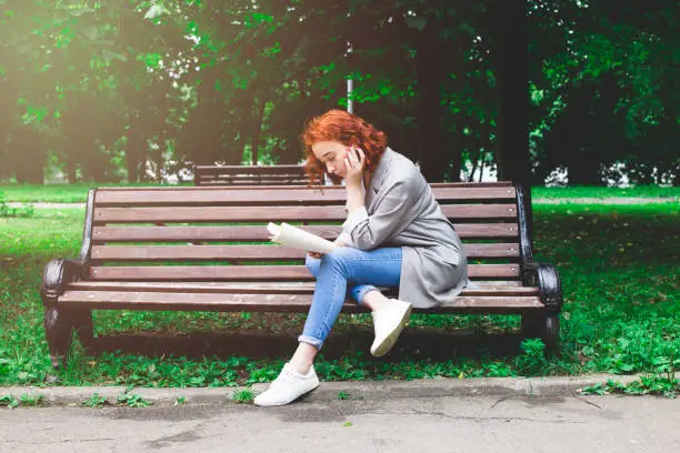 A girl is reading a book on a park bench. The girl has red hair and blue jeans