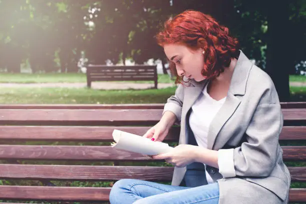 A girl is reading a book on a park bench. The girl has red hair.