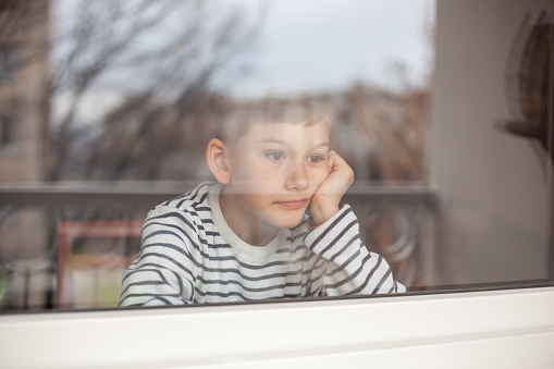 Blond boy of 7 years looking out a window. He seems a bit sad
