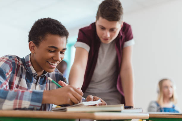 istock Smiling african american boy writing in textbook with classmate standing behind 1014150002