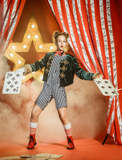 The nice young girl dressed in a magician costume shows the circus performance. The preadolescent girl is surprised looking at the camera. She is holding the large playing cards in her hands. Studio shooting on red background with glowing star and the striped theatrical curtain