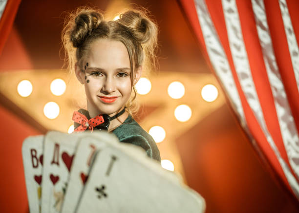 The nice young girl dressed in a magician costume shows the circus performance. The preadolescent girl is smiling looking at the camera. The large playing cards on foreground. Studio shooting on red background with glowing star and the striped theatrical curtain