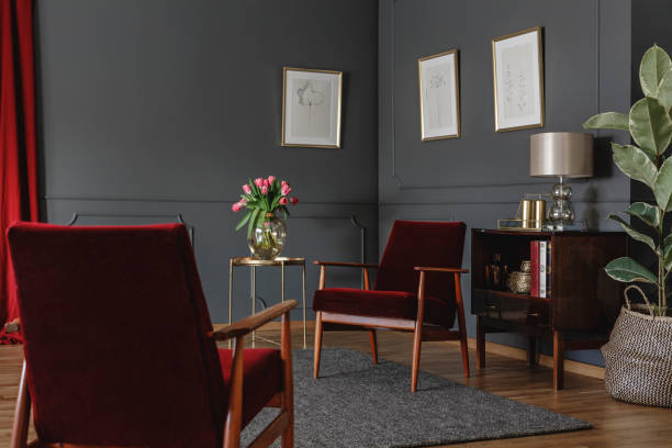 istock Two red armchairs standing in grey sitting room interior with minimal posters on walls with molding, wooden cupboard with lamp and decor and gold metal table with fresh tulips in vase 1014167632