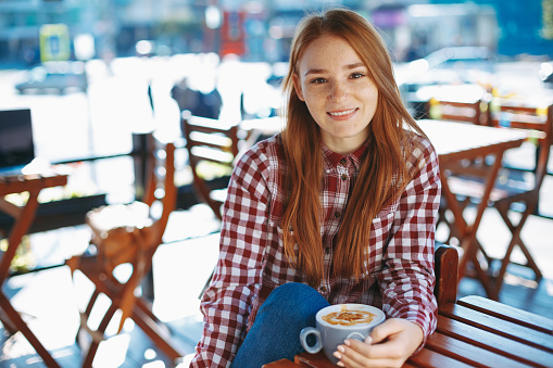 Coffee concept. Portrait of attractive young woman drinking coffee outdoors. Natural reddish girl with freckles, dressed in plaid shirt, smiling while holding a big cup of coffee latte.