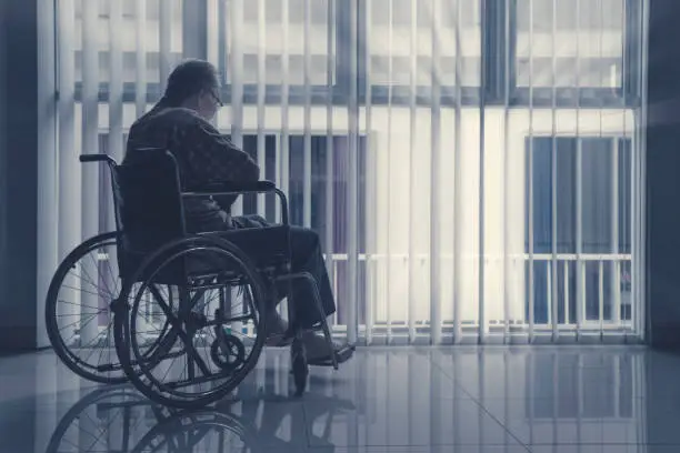 Lonely elderly man sitting on wheelchair near the window at home