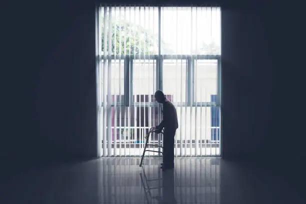 Silhouette of lonely old man walking near the window with a walker