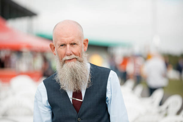 A headshot of a older man looking at the camera who is standing outdoors. He is wearing a navy waistcoat with a blue shirt and a red tie.