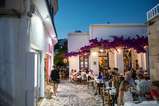 Milos island, Greece - June 11, 2018: Sunset scene in Plaka village which is full of shops and restaurants. People are dining in one of the restaurants decorated with bougainvillea flowers.