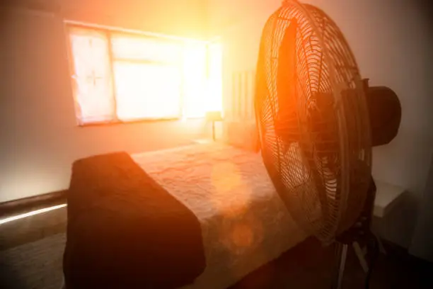 Electric fan next to bed with sunshine coming through the window.