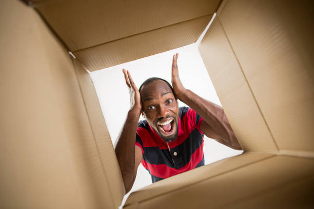 The surprised afrocan man unpacking, opening carton box and looking inside. The package, delivery, surprise, gift lifestyle concept. Human emotions and facial expressions concepts