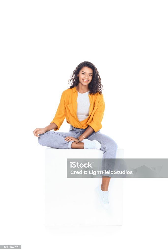 teenage african american student girl sitting on white cube and looking at camera isolated on white Sitting Stock Photo