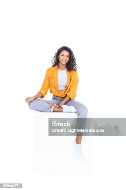 Teenage African American Student Girl Sitting On White Cube And Looking At Camera Isolated On White Stock Photo - Download Image Now