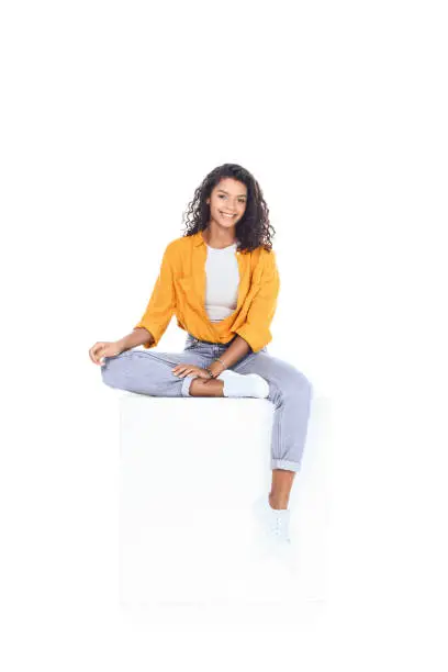 Photo of teenage african american student girl sitting on white cube and looking at camera isolated on white