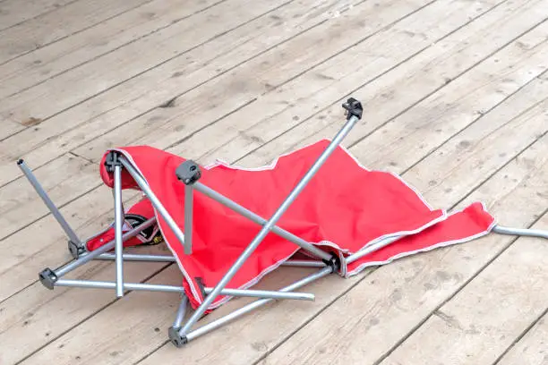 A red fabric broken beach or lawn chair on a wooden deck. The metal legs are broken and in disarray.