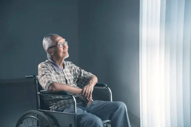 Elderly man looks happy while sitting in the wheelchair and daydreaming by the window. Shot in the retirement home