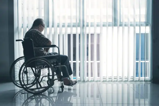 Picture of sad elderly man sitting in the wheelchair while looking out the window in the retirement home