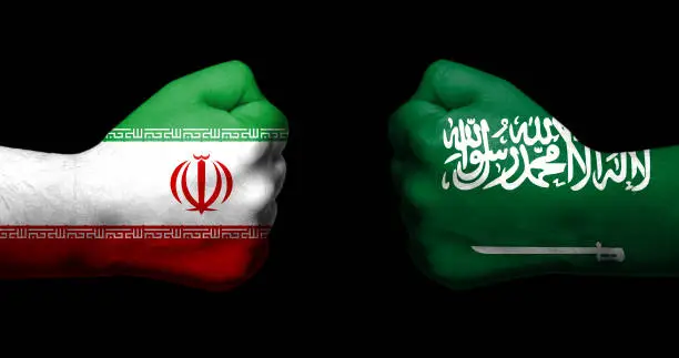 Flags of Iran and Saudi Arabia painted on two clenched fists facing each other on blue background/Iran - Saudi Arabia conflict concept