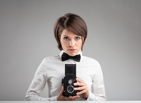 Stylish vintage female photographer wearing a bow tie holding her camera to her chest as she composes her image with a serious expression over grey