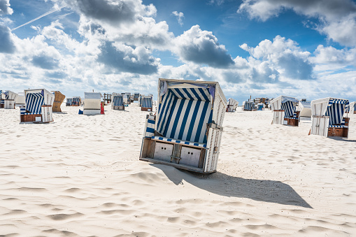 Typical roofed wicker beach chairs in blue-white stripes on white sandy german north sea beach under beautiful blue summer skyscape at the beach of St. Peter-Ording, Nordfriesland, Germany.