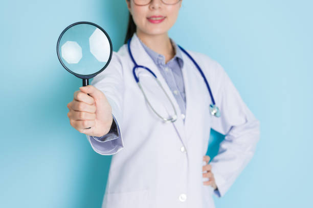 smiling young doctor standing in blue background smiling young female doctor standing in blue background and face to camera showing magnifier tool. photographic enlarger stock pictures, royalty-free photos & images