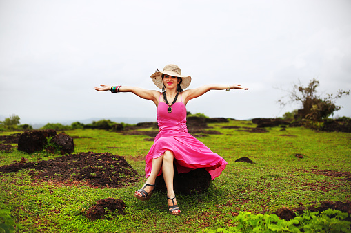 Young Indian fashion girl wearing pink dress and wearing hat, she sitting on a rock and doing hand raised freedom concept outdoors nature.