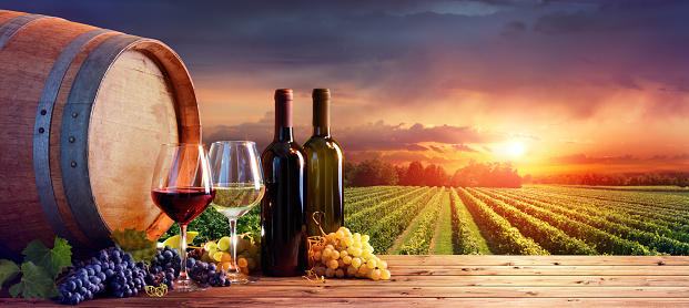 Bottles And Glasses With Ripe Grapes And Wooden Barrel At Sunset