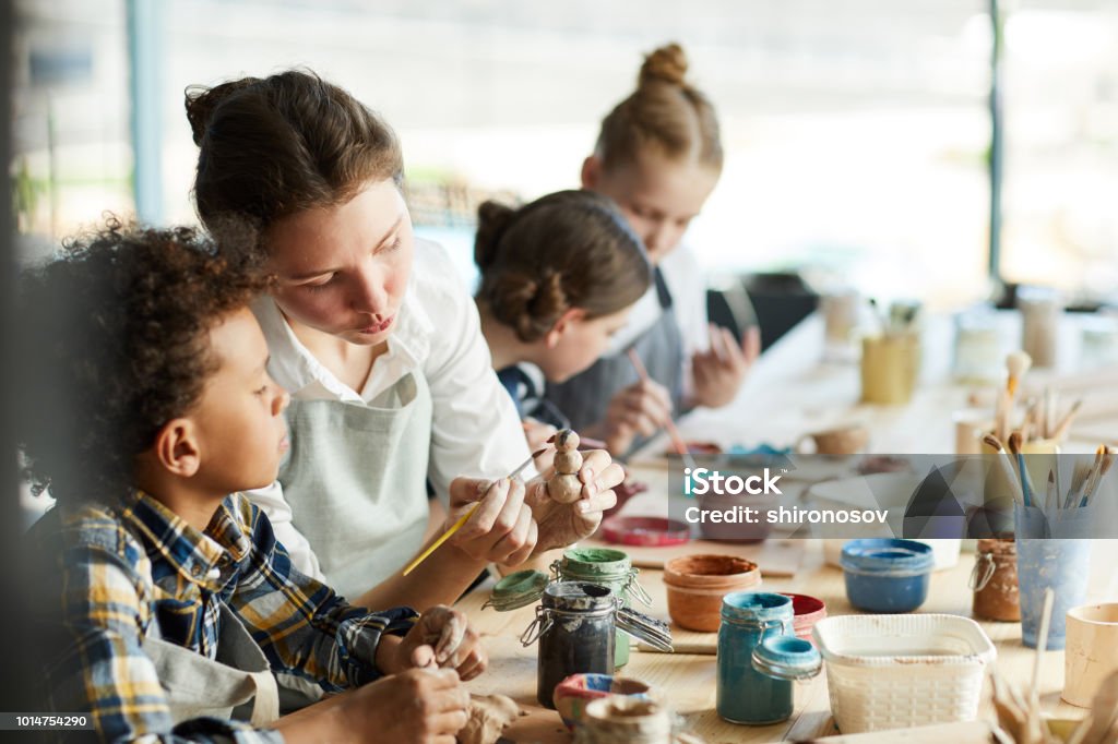 Advice of teacher Young teacher showing one of pupils clay item and giving advice about painting it Child Stock Photo