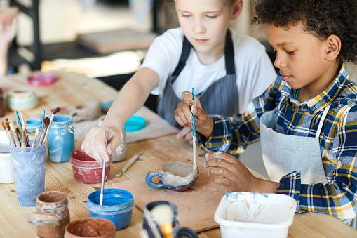 Two youthful claaamates sitting by table and painting self-made clay items at lesson