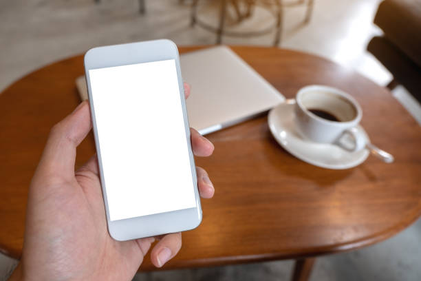 istock Mockup image of hand holding white mobile phone with blank desktop screen with coffee cup and laptop on table 1014758654