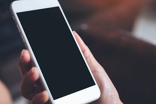 Mockup image of a hand holding white mobile phone with blank black desktop screen with blur background