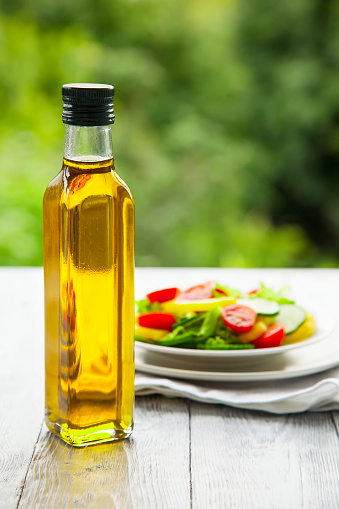 glass bottle of olive oil, focus on her.Background plate with vegetable salad on natural background on wooden table