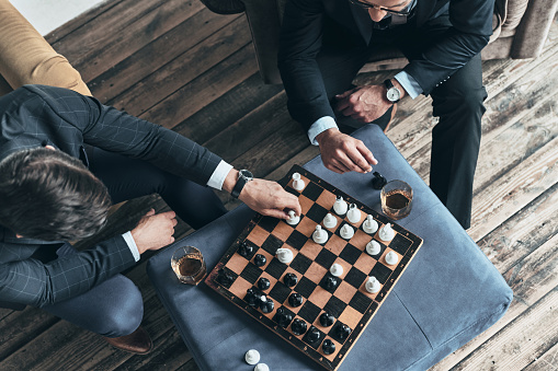 Top view of young thoughtful men in full suits playing chess while sitting indoors