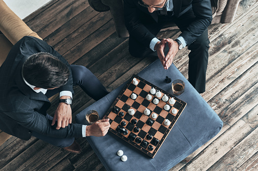 Top view of young thoughtful men in full suits playing chess while sitting indoors