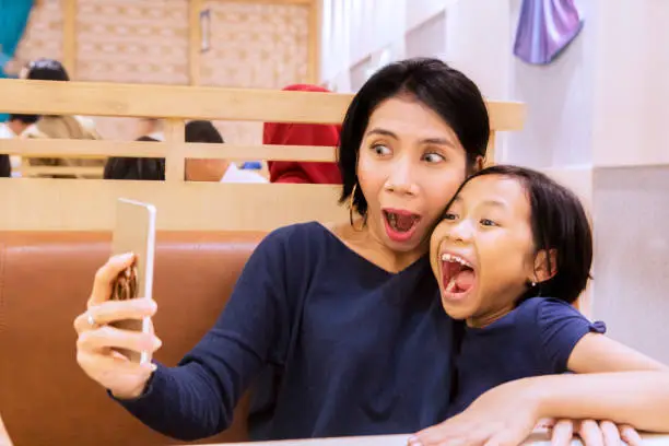 Picture of a young woman with her daughter using a smartphone while taking funny selfie in the restaurant