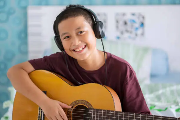 Image of Asian little boy wearing headset while playing acoustic guitar. Shot at home
