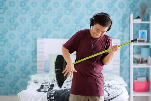 Picture of a cute little boy wearing headset while playing guitar by using broom in the bedroom