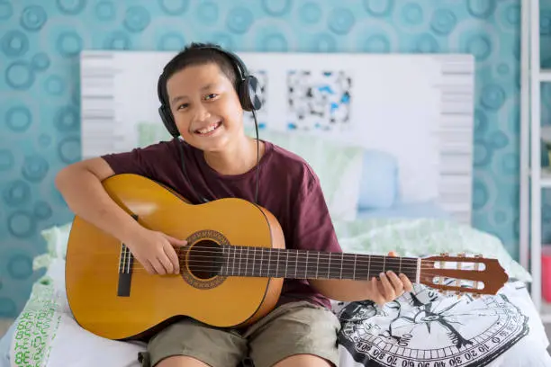 Image of Asian little boy wearing headset while practicing to play acoustic guitar in the bedroom
