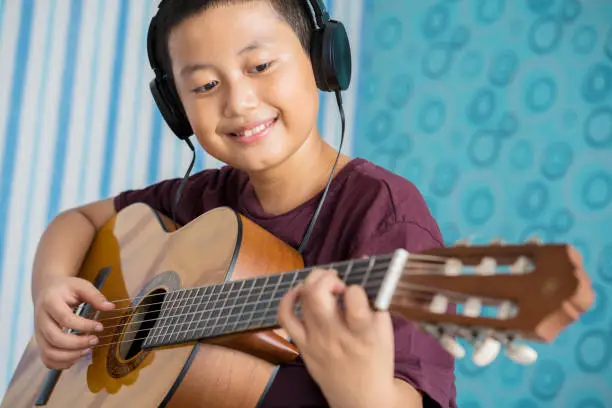 Closeup of happy little boy practicing to play acoustic guitar while wearing headset. Shot in the bedroom