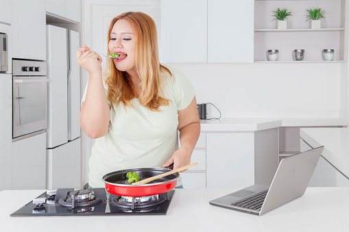 Picture of beautiful fat woman tasting food while standing in the kitchen