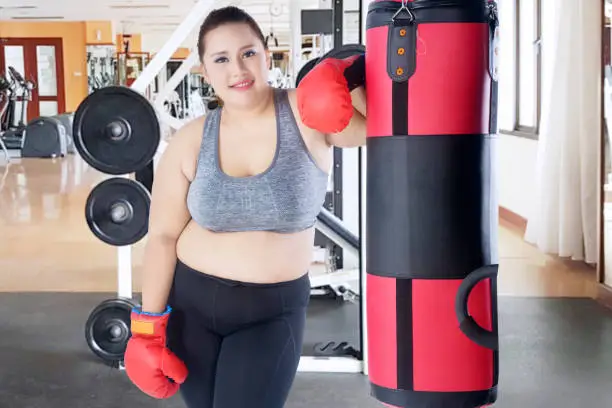 Portrait of Caucasian fat woman smiling at the camera while leaning on a bag boxing, isolated on white background