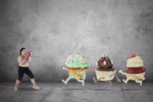 Image of Caucasian fat woman ready to fight with tasty cupcakes while wearing boxing gloves. Concept of diet