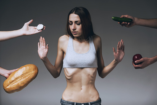 Lean Woman Refuses to Eat. Anorexia Concept. Thin Girl in Center Rejects. Food Offered from Different Sides. Belly Ribs. Isolated Background. Skinny Waist. Abnormal Weight Loss. Unhealthy Nutrition.