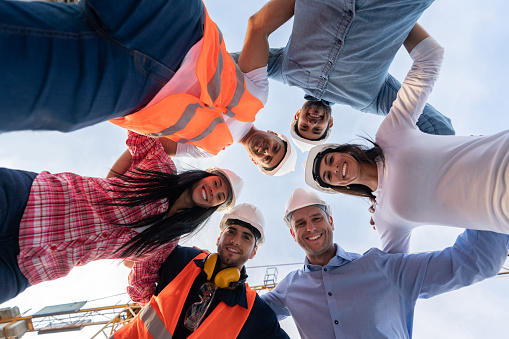 Happy team of engineers hugging in a circle at a construction site smiling and wearing helmets