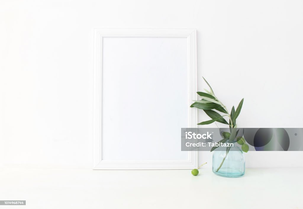 White vertical blank wooden frame mockup with a green olive branches in blue glass vase lying on the table. Mediterranean summer poster product design. Styled stock feminine photography. Home decor. White vertical blank wooden frame mockup with a green olive branches in blue glass vase lying on the table. Mediterranean summer poster product design, styled stock feminine photography. Home decor. Picture Frame Stock Photo